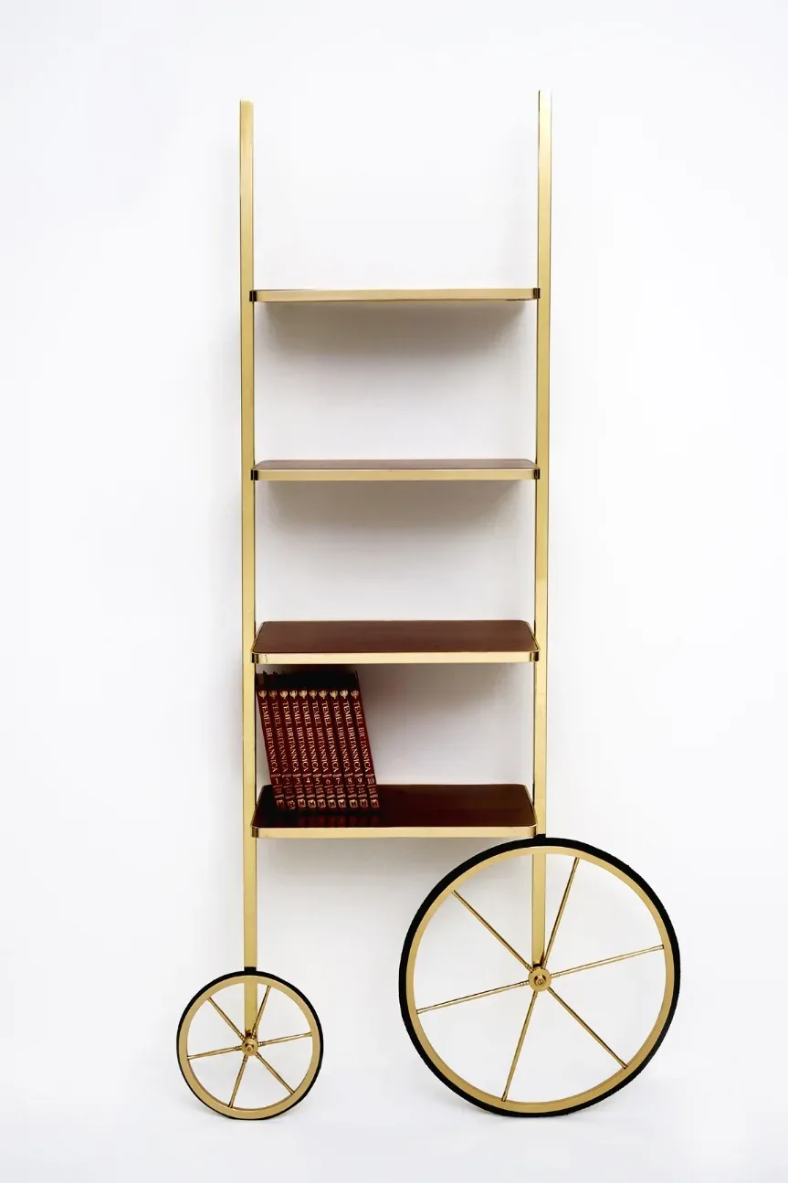 Brass and wood shelving unit with four shelves and two decorative wheels, displayed against a white wall by Merve Kahraman.