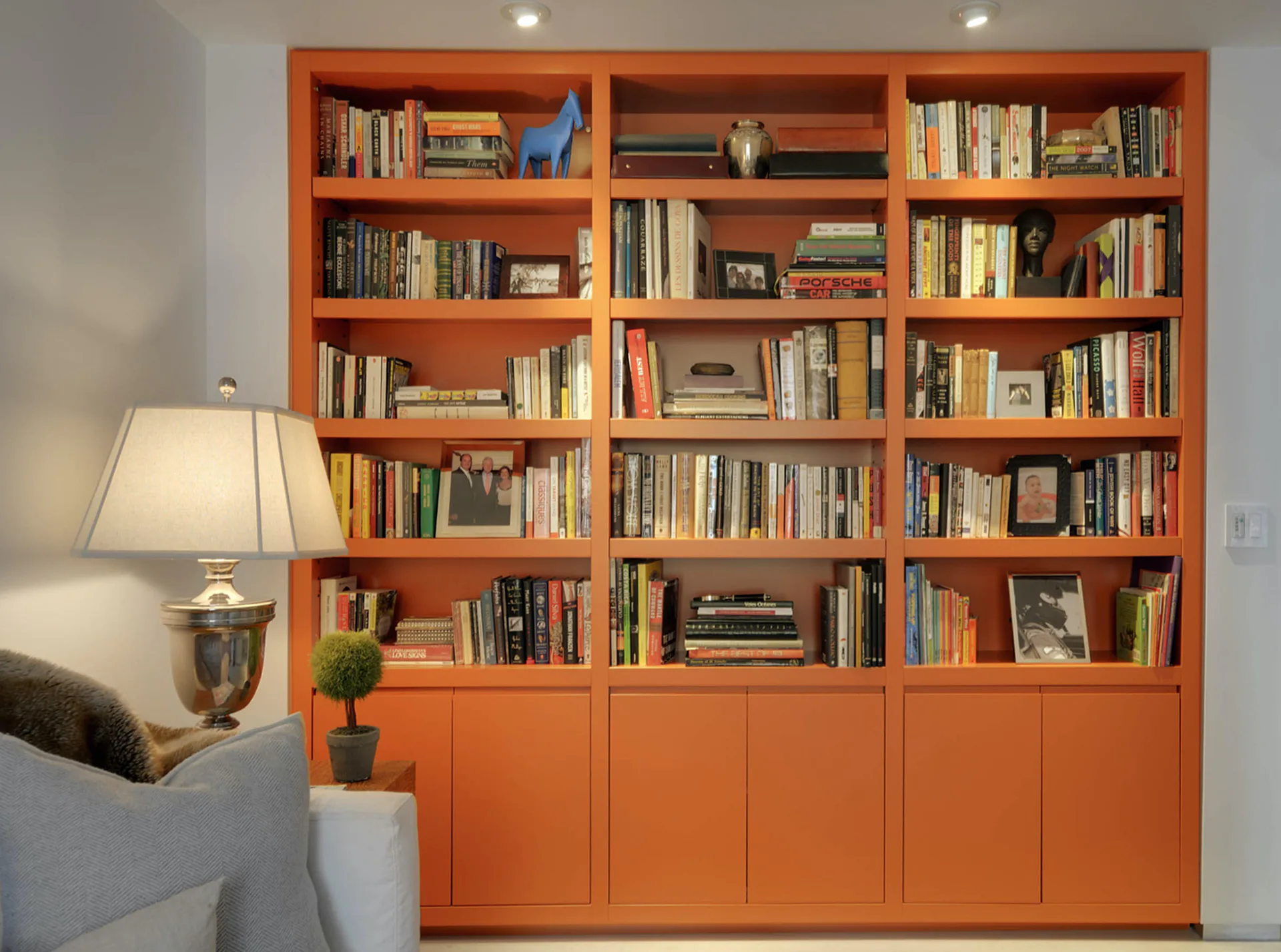 Bright residential interior featuring a floor-to-ceiling orange custom bookcase with open shelving above and closed cabinetry below, filled with books and decorative objects, beside a gray armchair and table lamp by Meryl Scheinman.