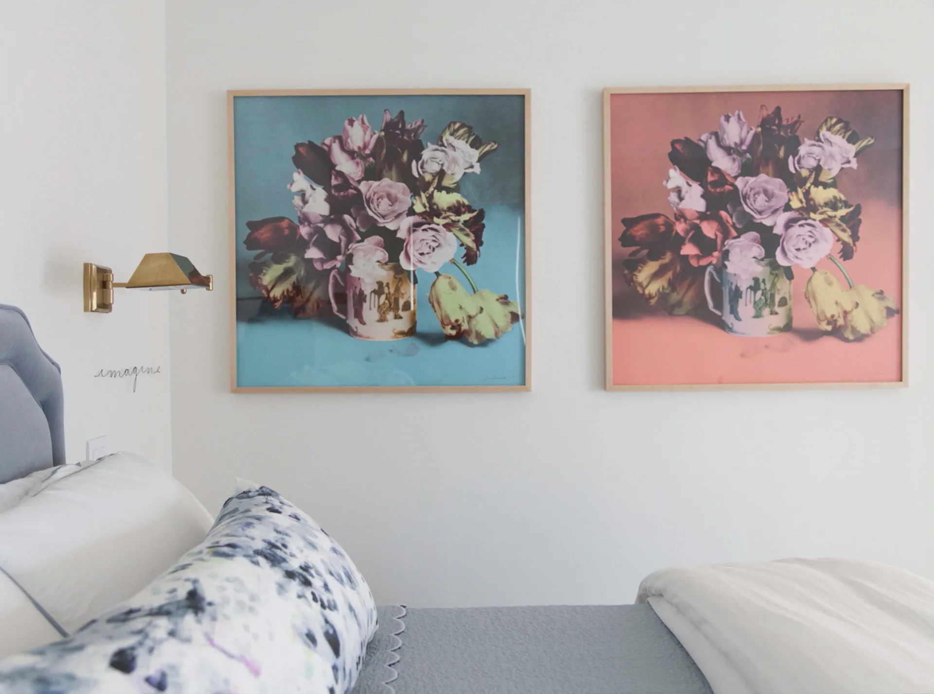 Minimalist bedroom interior featuring two framed floral artworks with teal and coral backgrounds, paired brass wall sconce, and white bedding by Meryl Scheinman.