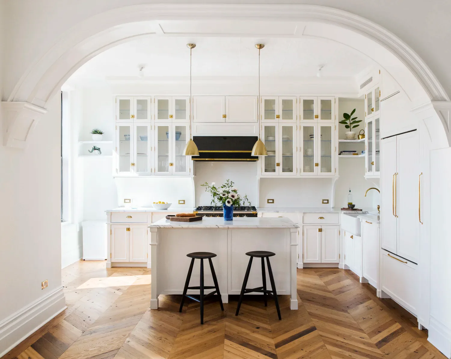 Bright white kitchen with cream cabinetry, island seating, herringbone wood flooring, and black pendant stools by Mia Jordan.