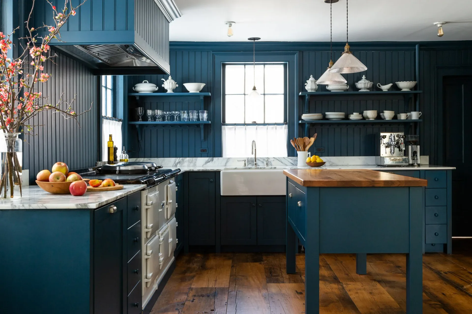 A chef's kitchen featuring deep teal cabinetry with open shelving, stainless steel farmhouse sink, wood countertops, and hardwood flooring, designed by Mia Jordan.