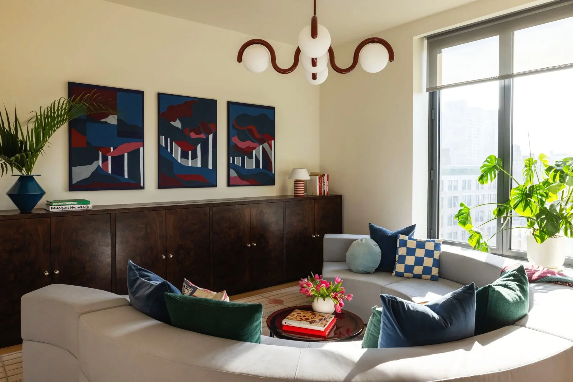 Modern living room with curved seating, dark wood cabinetry, geometric artwork, and abundant natural light from large windows, designed by Mia Jordan.