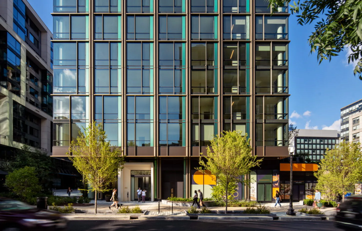 Modern mixed-use building facade with dark bronze metalwork, expansive glazing, and ground-floor retail activation on a tree-lined urban street by Michael Arad.