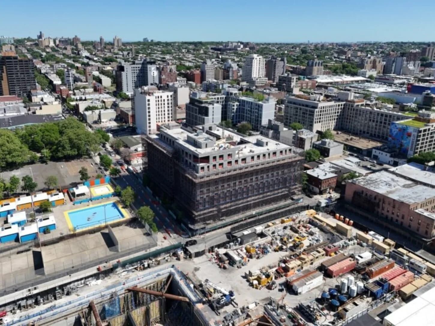 Aerial view of an urban waterfront development with construction materials and equipment visible, overlooking a city neighborhood with residential and commercial buildings by Michael Arad.