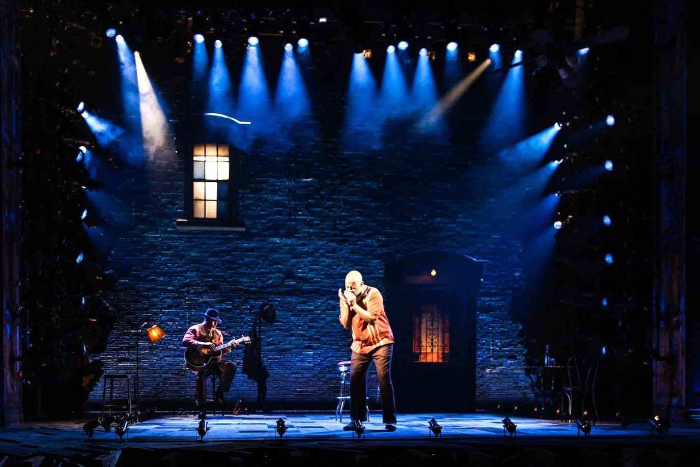 Theater stage set featuring a brick wall backdrop with illuminated windows, blue atmospheric lighting, and performers on stage, designed by Michael Carnahan.