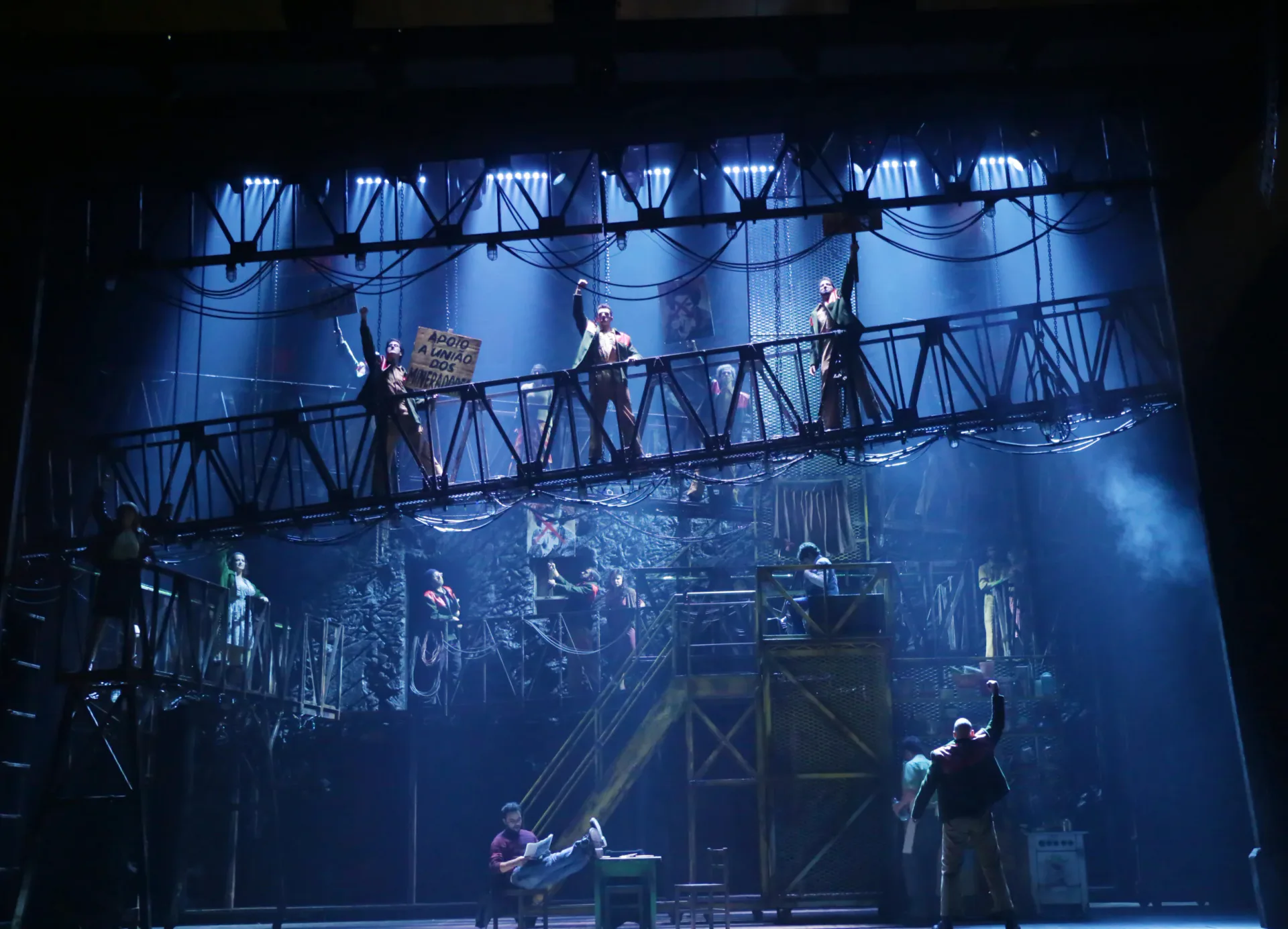 Theatrical stage set featuring multiple metal bridge structures and industrial framework in blue lighting, designed by Michael Carnahan.