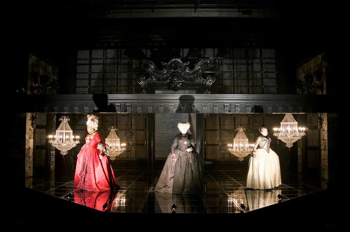 Elegant retail window display featuring three mannequins in formal gowns illuminated by crystal chandeliers against dark architectural backdrop, by Michael Carnahan.