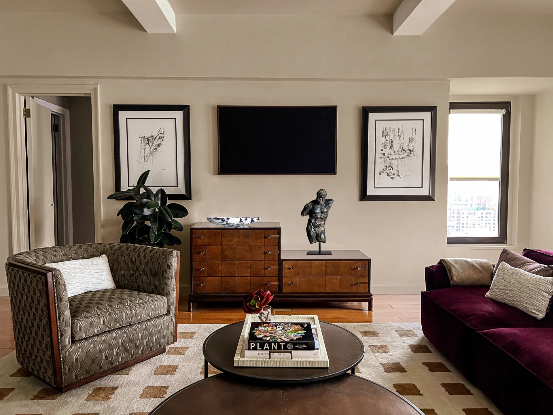 Modern living room with neutral palette, patterned armchair, burgundy sofa, wooden mid-century dressers, round coffee table, and wall-mounted TV by Michael Gilbride.