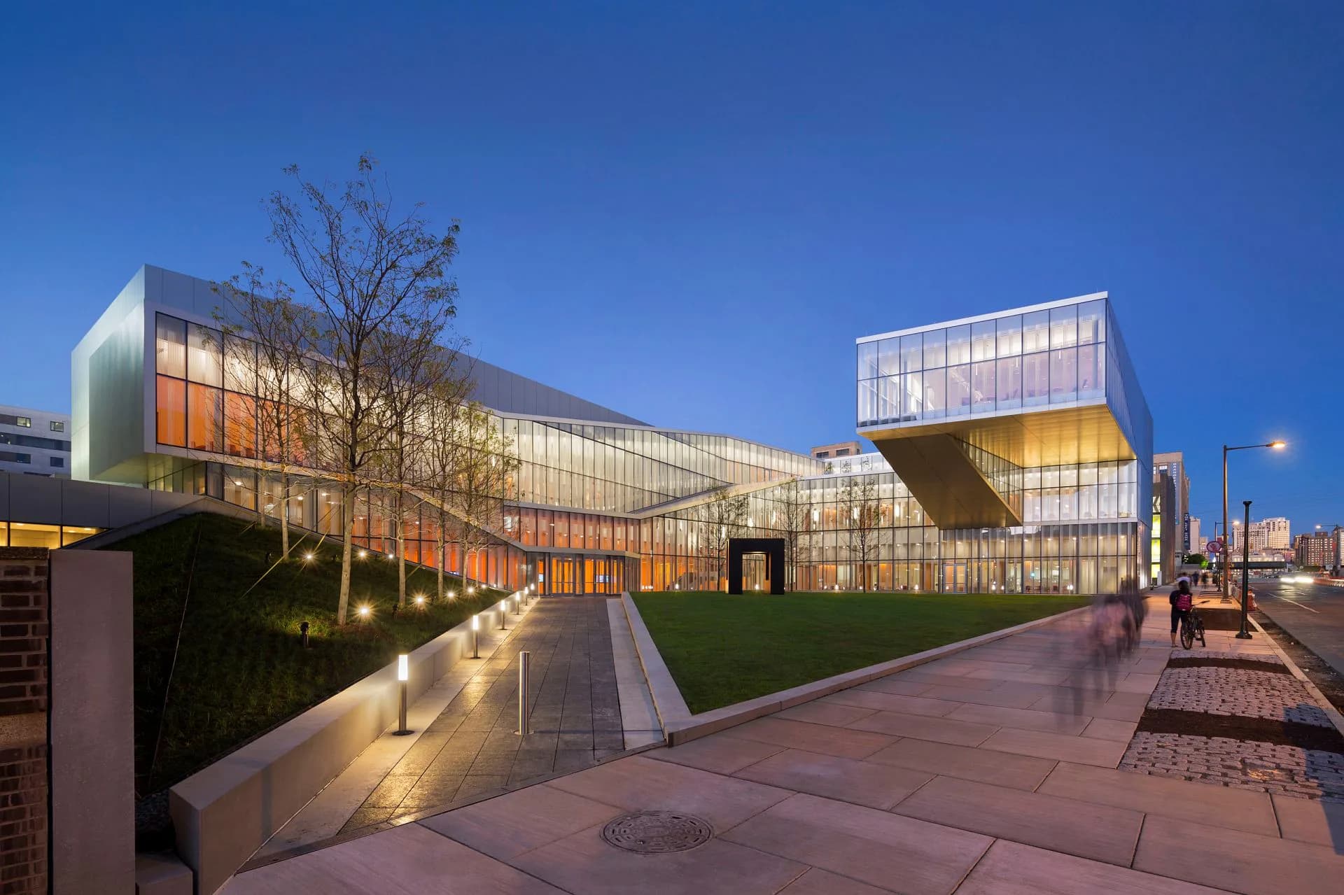 Modern institutional building with glass and metal facades illuminated at dusk, featuring angular cantilevers and a contemporary plaza design by Michael Manfredi.