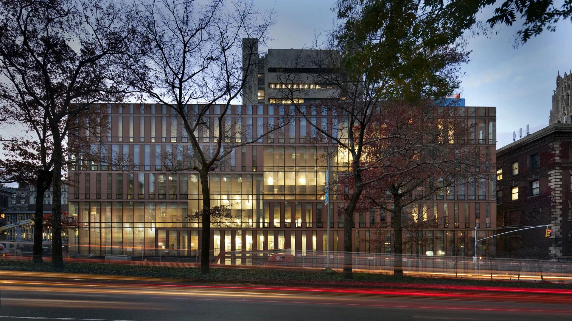 Modern institutional building with glass and brick facade illuminated at dusk, featuring vertical window patterns and contemporary architectural design by Michael Manfredi.