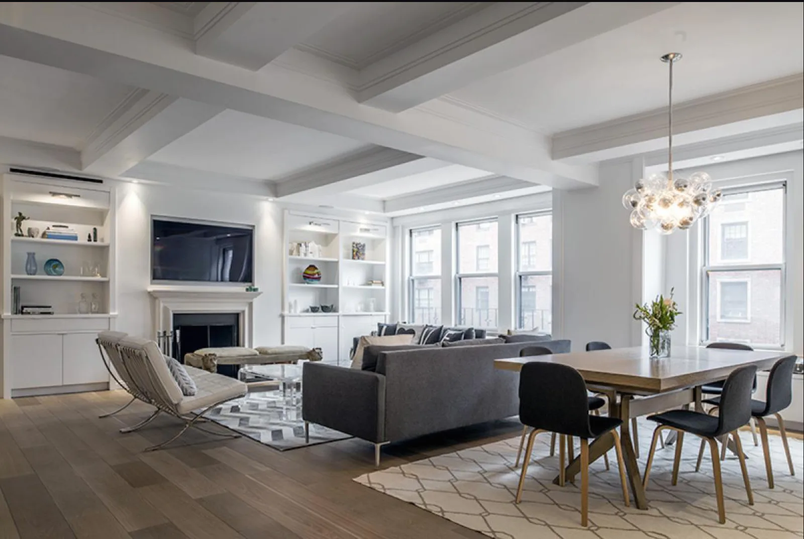 Modern open-plan living and dining space with coffered ceiling, dark sectional sofa, fireplace with white built-ins, wood dining table, pendant lighting, and large windows by Michal Mufson.
