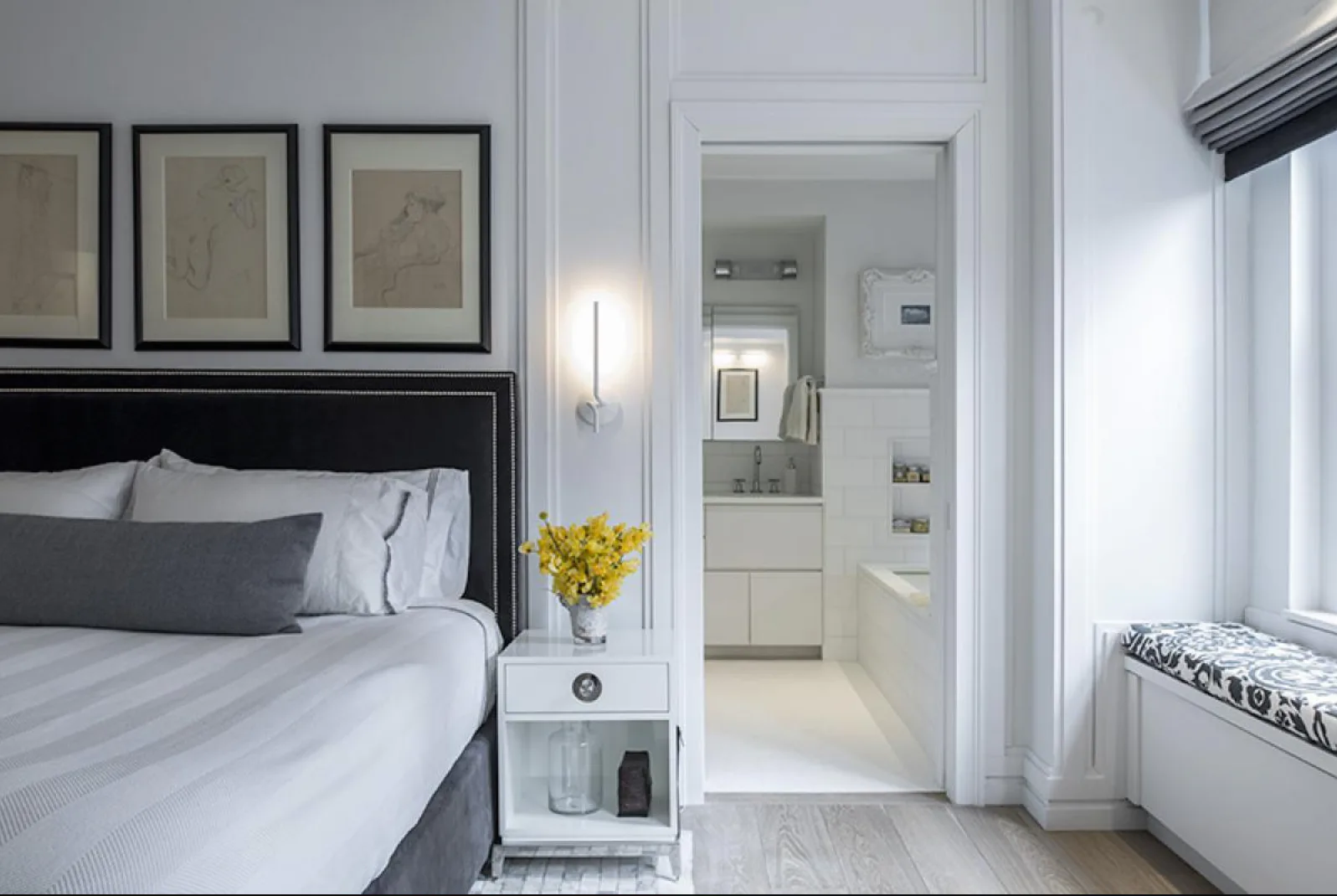 Modern bedroom with black-framed bed, white bedding, three framed artworks, ensuite bathroom visible through doorway, bright natural light, designed by Michal Mufson.