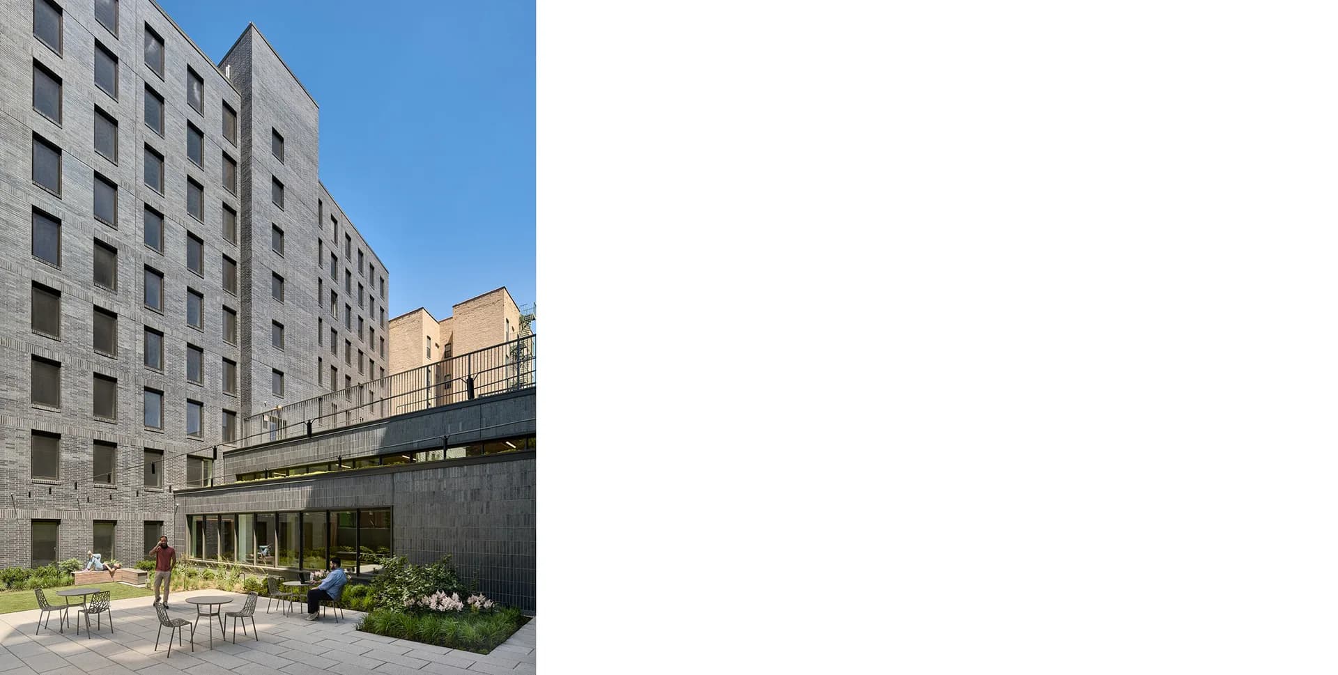 Contemporary mixed-use building exterior with residential tower and ground-level terrace featuring outdoor seating, landscaping, and dark architectural elements by Michelle Romano.