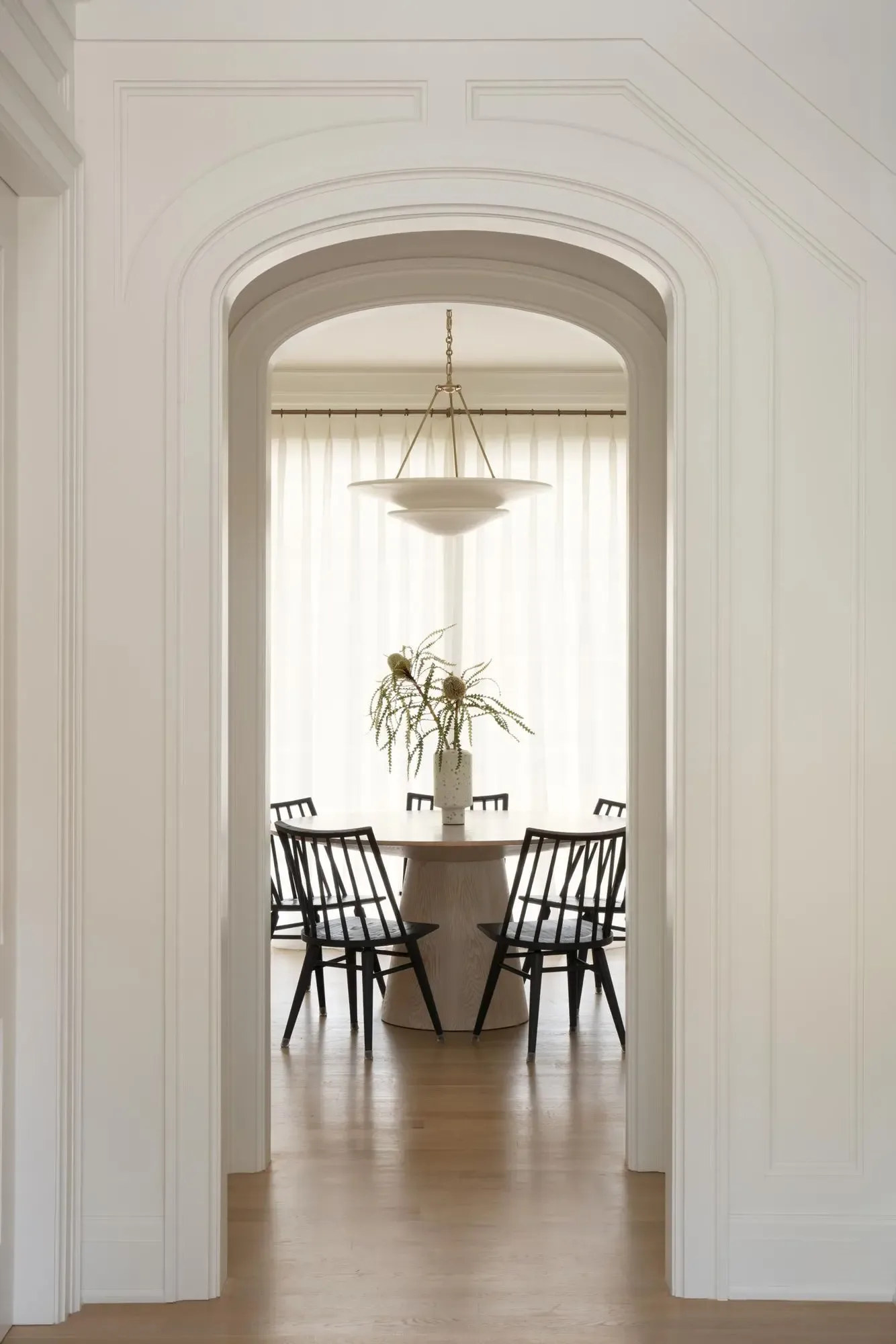 Elegant dining room with arched doorway, neutral palette, brass pendant light, and black spindle chairs around a round table by Michelle Waugh.