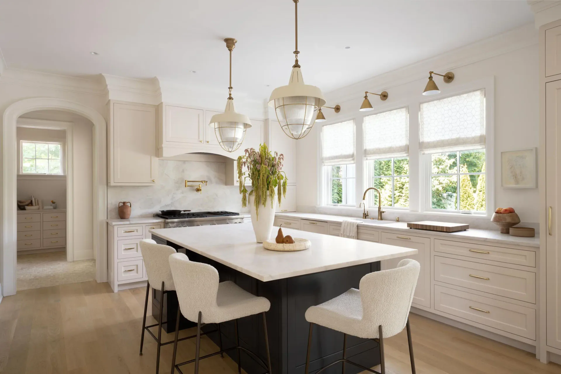 Bright white kitchen with black island, cream upholstered seating, pendant lighting, and large windows overlooking greenery by Michelle Waugh.