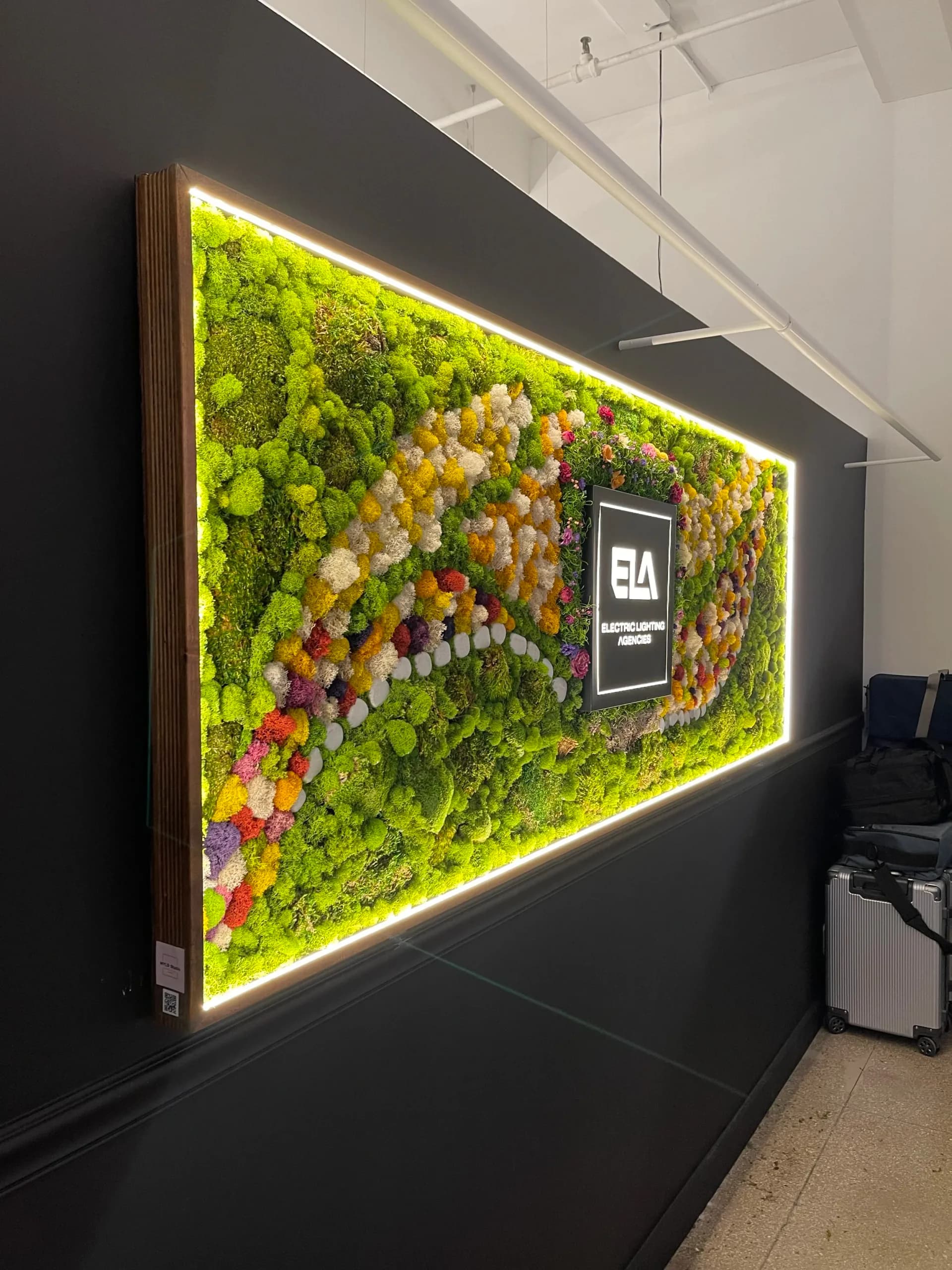 Modern interior installation featuring a large illuminated moss wall with colorful preserved flowers and green moss panels, framed with neon lighting, by Michelle Yang.