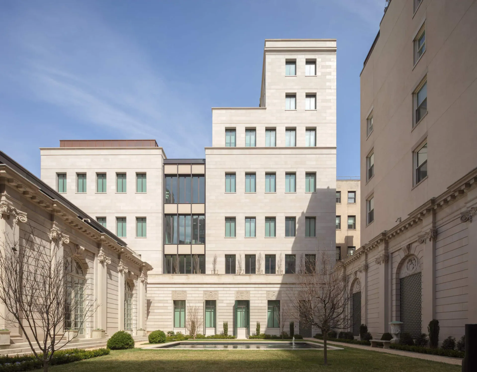 Modernist courtyard with cream-colored stepped buildings, manicured landscaping with shrubs and bare trees, and classical stone facades flanking the contemporary architecture by Michi Ushio.