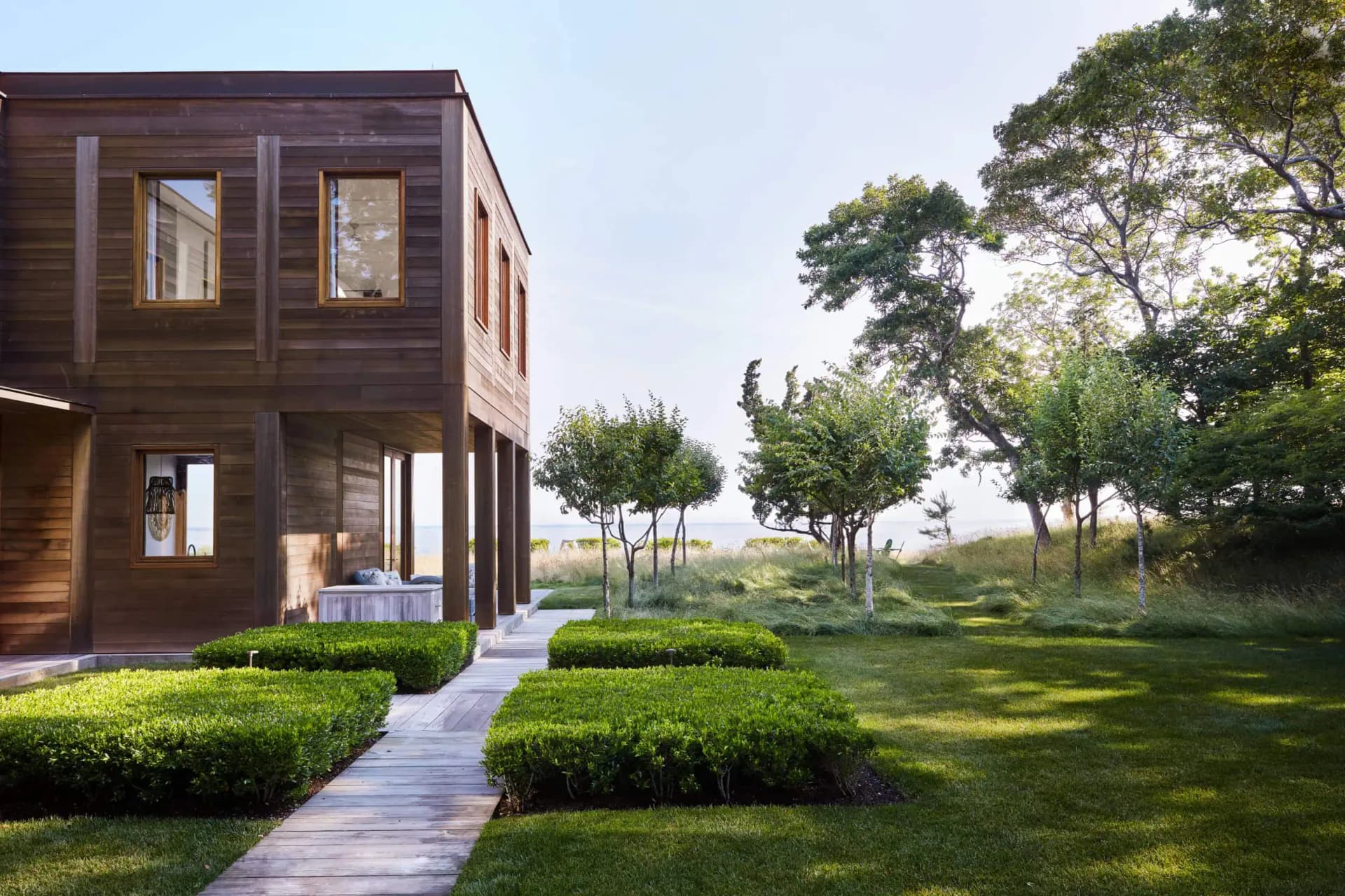 Modern wooden-clad residential structure with expansive windows overlooking manicured hedges and open landscape with mature trees, designed by Michi Ushio.