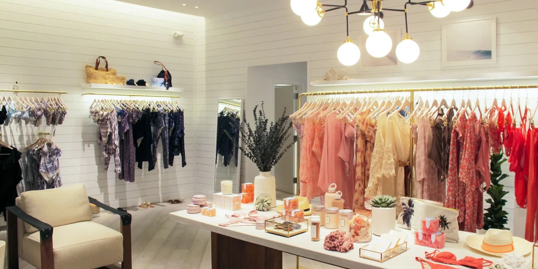 Bright retail clothing boutique with white brick walls, globe pendant lighting, coral and neutral-toned garments on display racks, and styled product table by Michiru Tanaka.