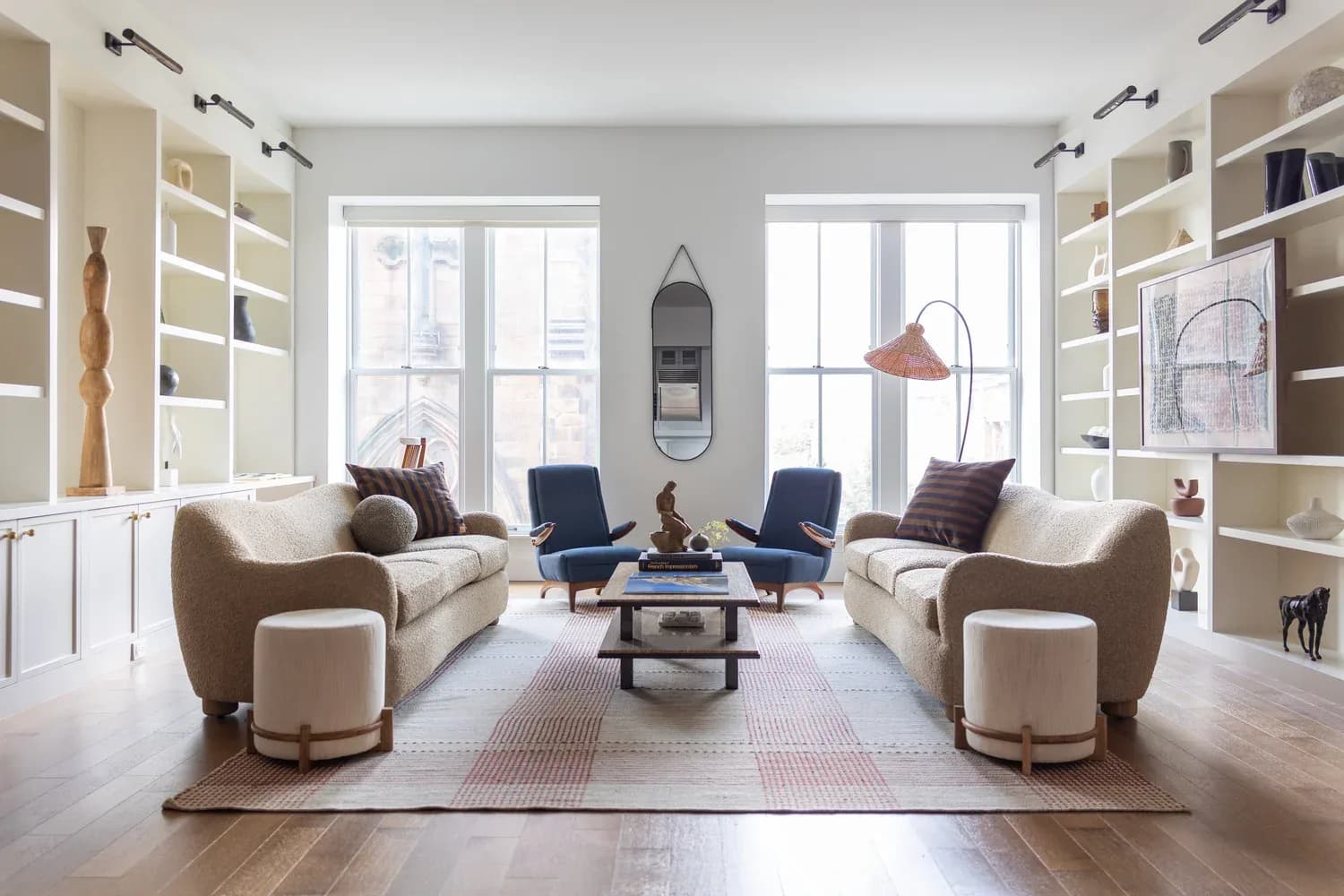 Modern living room with symmetrical seating arrangement, built-in shelving, large windows, and contemporary furnishings in neutral tones by Mika Lipson.