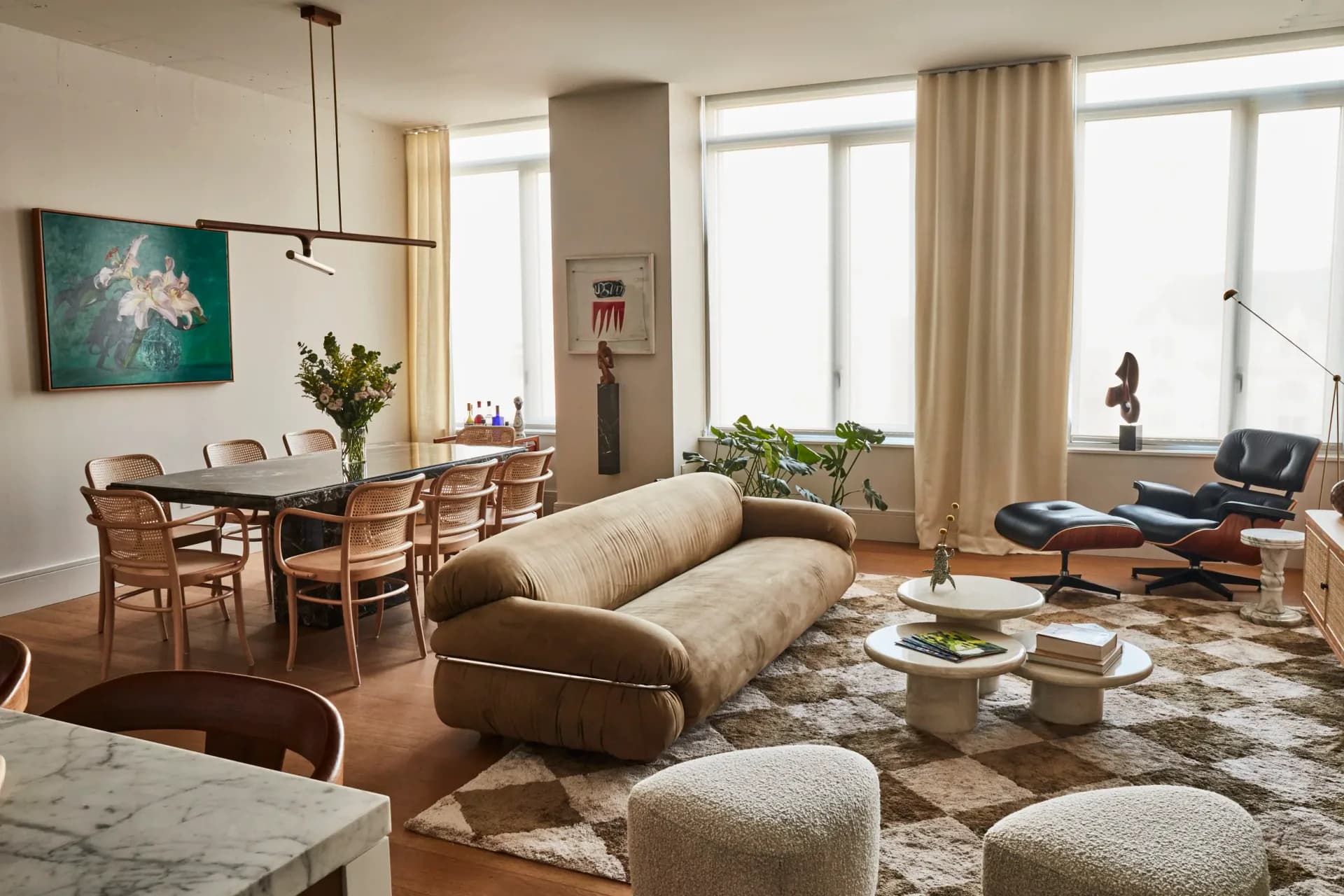 Modern living room with curved beige sofa, wooden dining area, large windows with cream curtains, patterned area rug, and lounge chair by Mika Lipson.