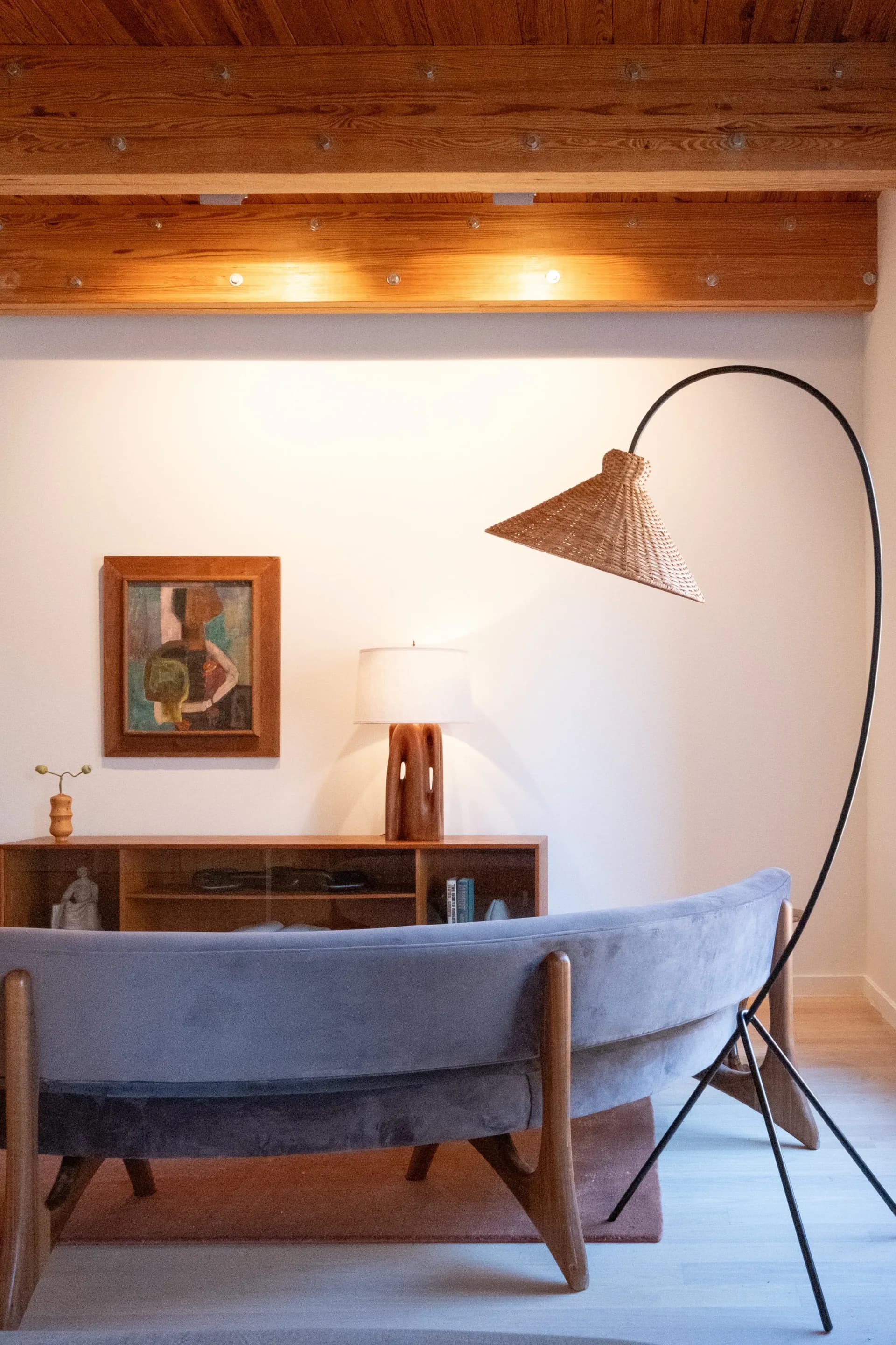Mid-century modern living room with blue upholstered sofa, wooden sideboard, arc floor lamp, framed artwork, and exposed wood ceiling with recessed lighting by Mika Lipson.