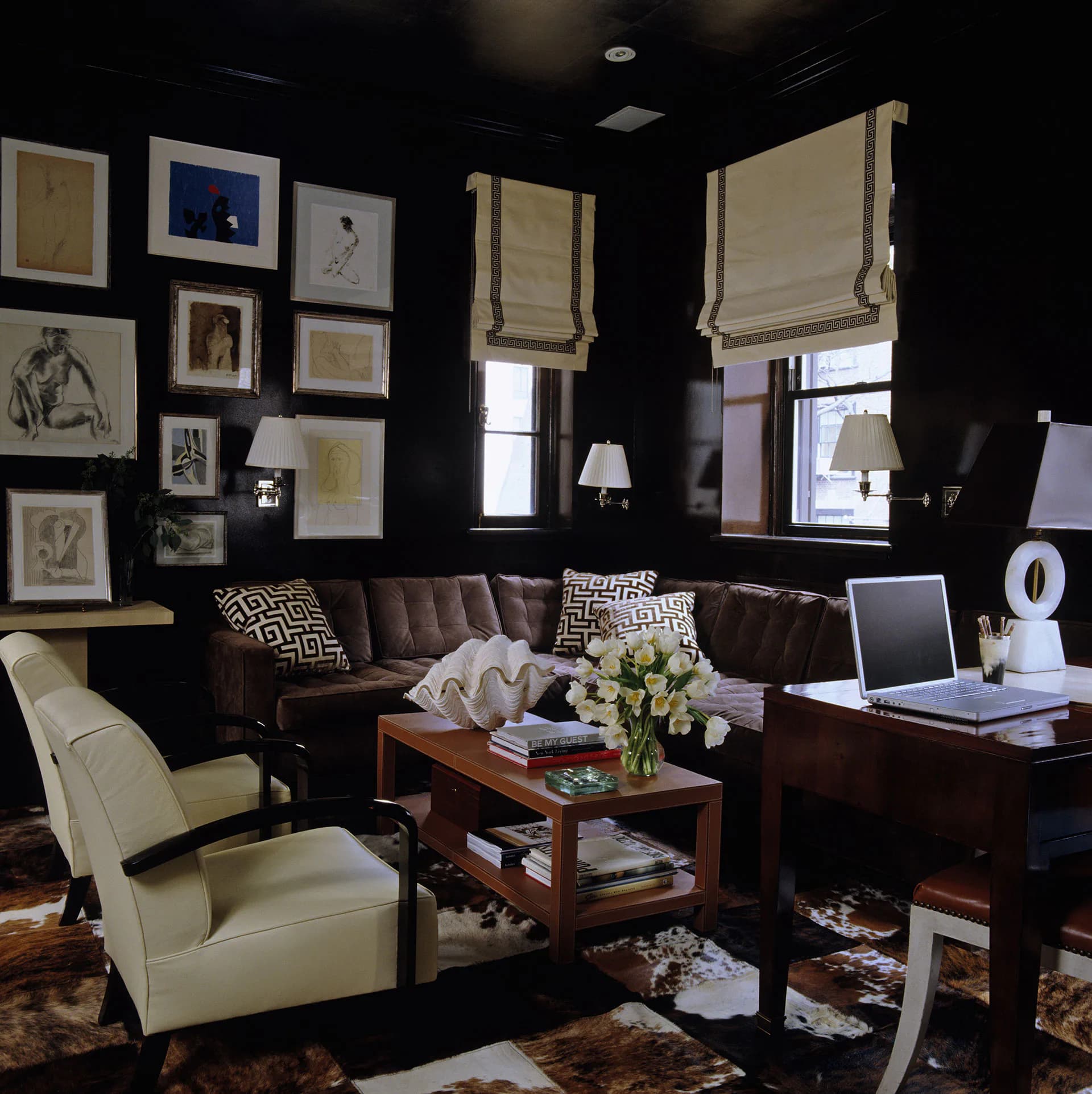 Sophisticated dark-walled study with chocolate leather sofa, cream armchair, wooden desk, gallery wall of framed artwork, and warm lighting by Miles Redd.