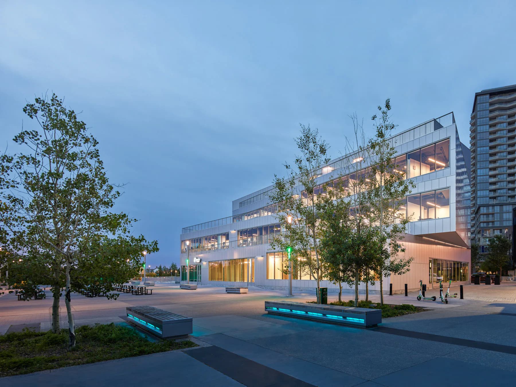 Modern institutional building with illuminated glass facades, horizontal layering, and integrated landscaping at dusk by Mimi Hoang.