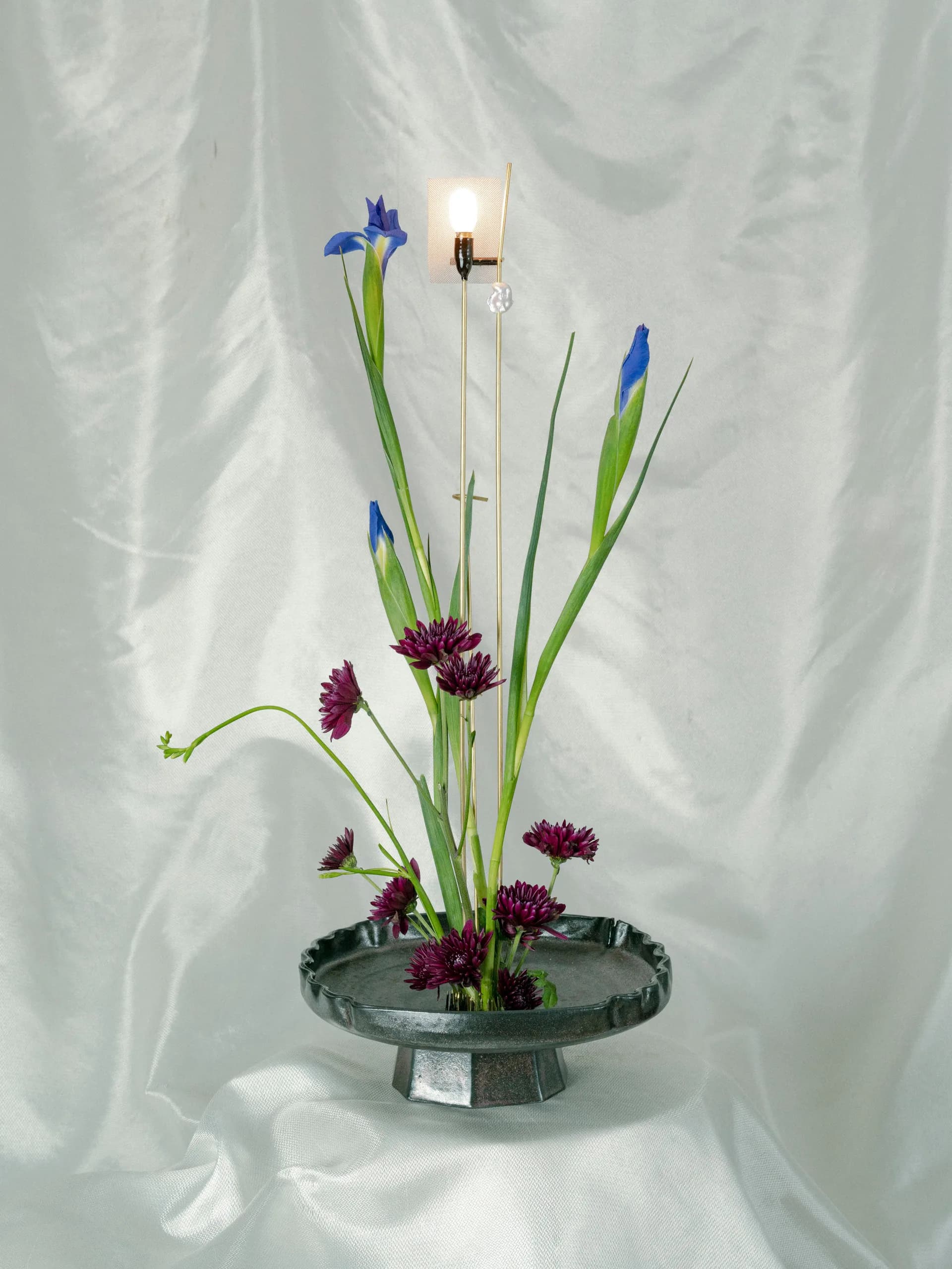 Minimalist floral arrangement featuring blue iris flowers, purple carnations, and green stems in a shallow gray pedestal vessel with a lit candle by Minjae Kim.