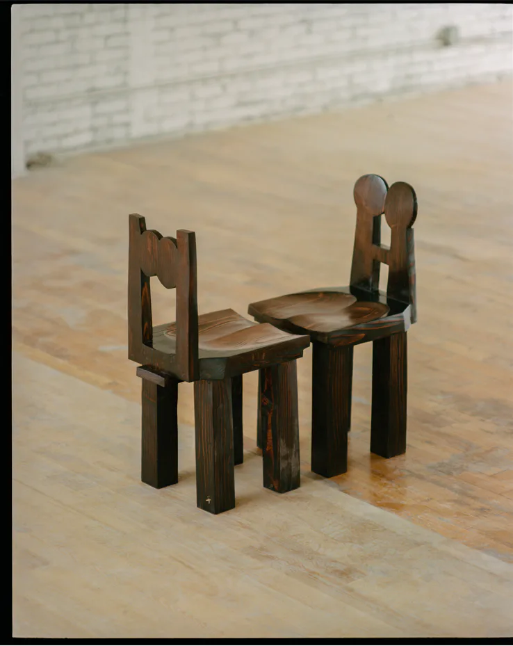 Two sculptural wooden chairs with organic cutout backrests sit on a polished wooden floor, demonstrating artistic furniture design by Minjae Kim.