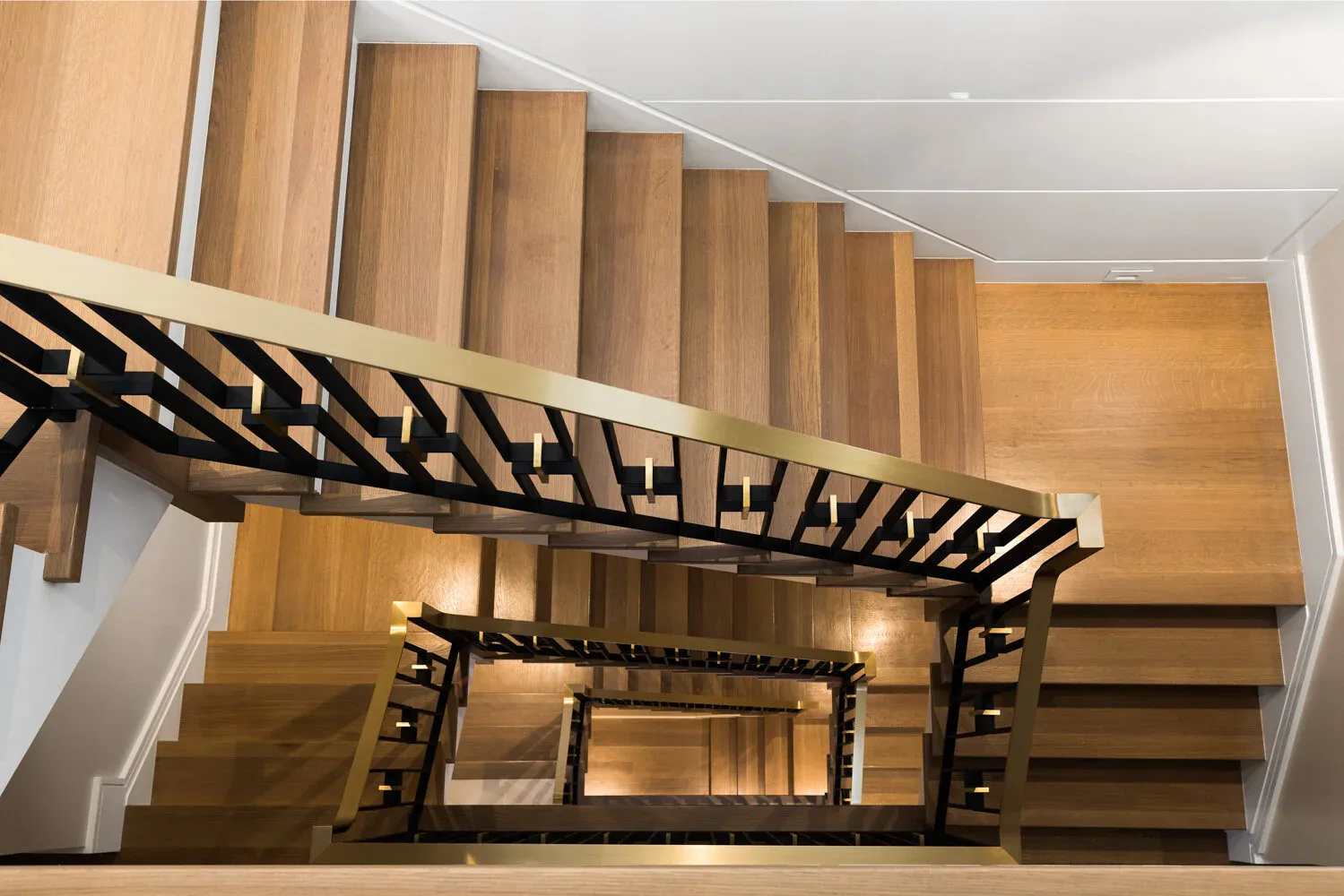 Modern wooden staircase with metal railings descends through a bright multi-level interior, showcasing warm wood tones and clean architectural lines by Miriam Lichtenstein.