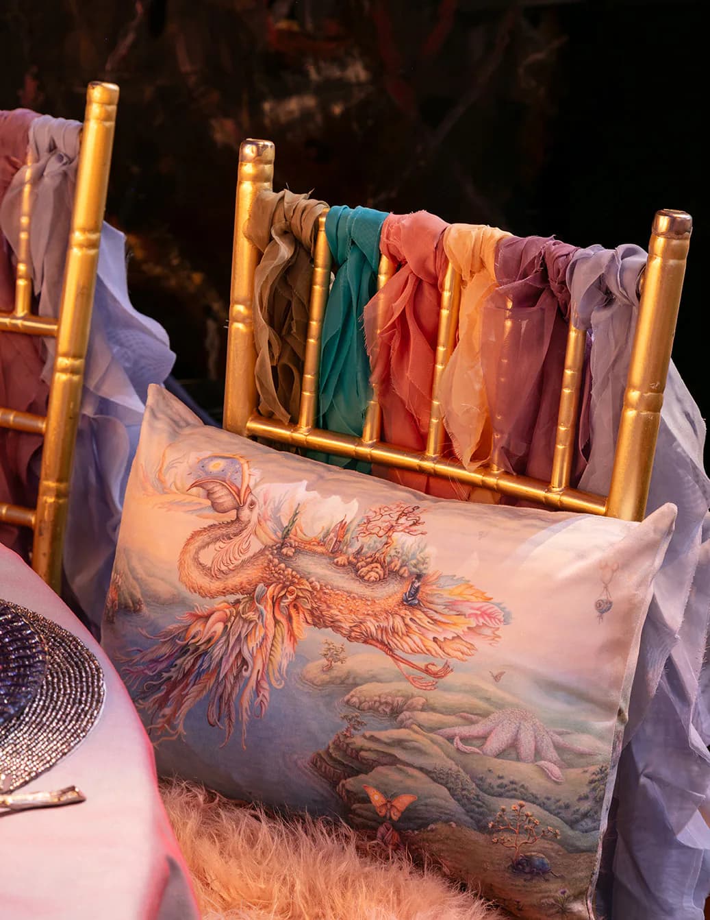 Colorful silk fabrics and textiles hang from a gilded bamboo rod beside a decorative pillow with mythical creature embroidery, showcasing textile design and styling work by Miriam Lissak.
