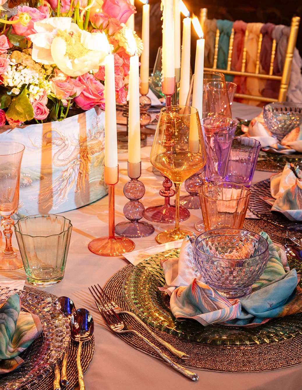 Elegant table setting with gold charger plates, crystal glassware, coral candlesticks, and pink floral centerpiece against warm lighting by Miriam Lissak.