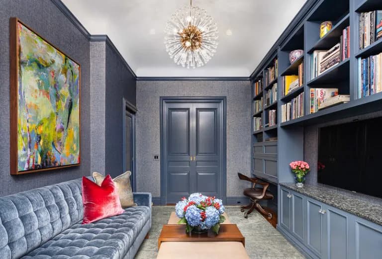 Contemporary navy library den featuring built-in bookshelves, quilted gray sofa with red accent pillow, brass starburst pendant, and brass-handled blue doors, designed by Miriam Lissak.