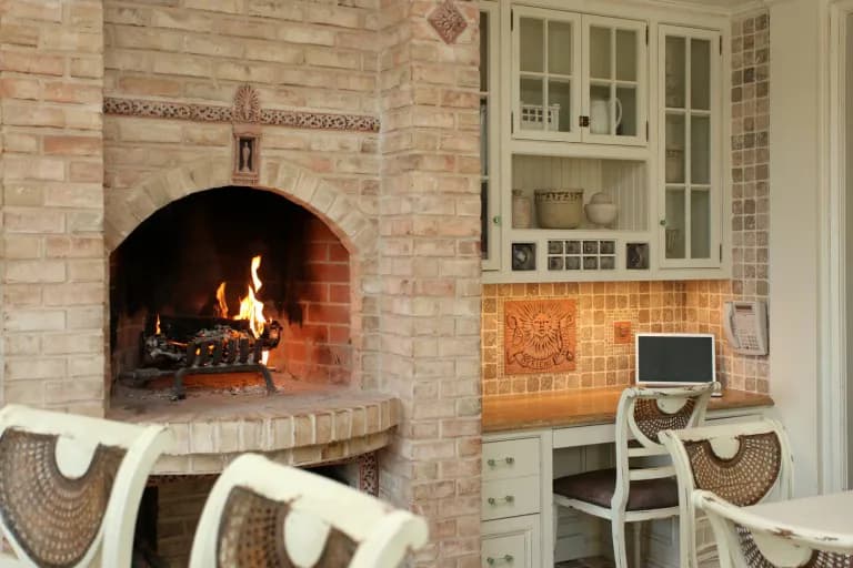 Brick kitchen and fireplace interior with arched hearth, white cabinetry, tiled backsplash, and workspace by Miriam Lissak.