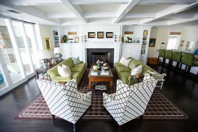 Symmetrical living room with coffered ceiling, green armchairs, fireplace, dark hardwood floors, and geometric area rugs by Mo Lebel.