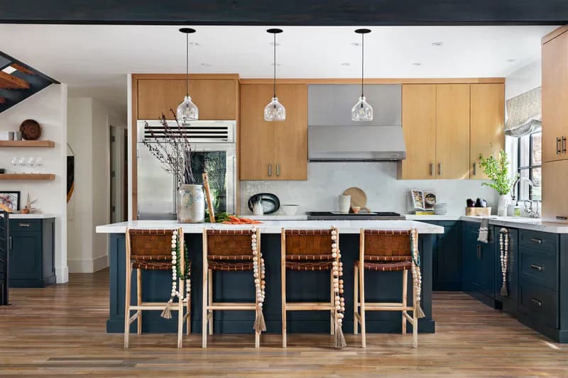 Modern kitchen with natural wood upper cabinets, navy lower cabinets, white waterfall island with woven bar stools, and pendant lighting by Mo Lebel.