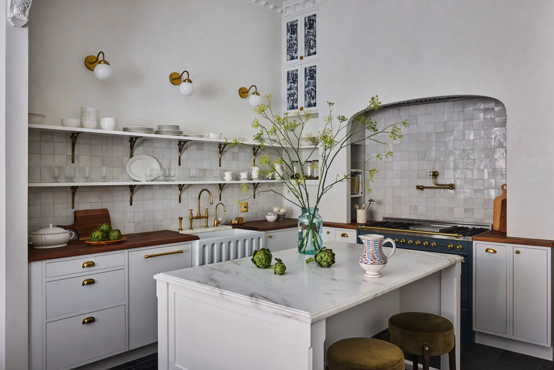 Modern white kitchen with marble countertops, brass fixtures, open shelving, and a central island with green stools, featuring soft natural lighting by Molly Portnof.
