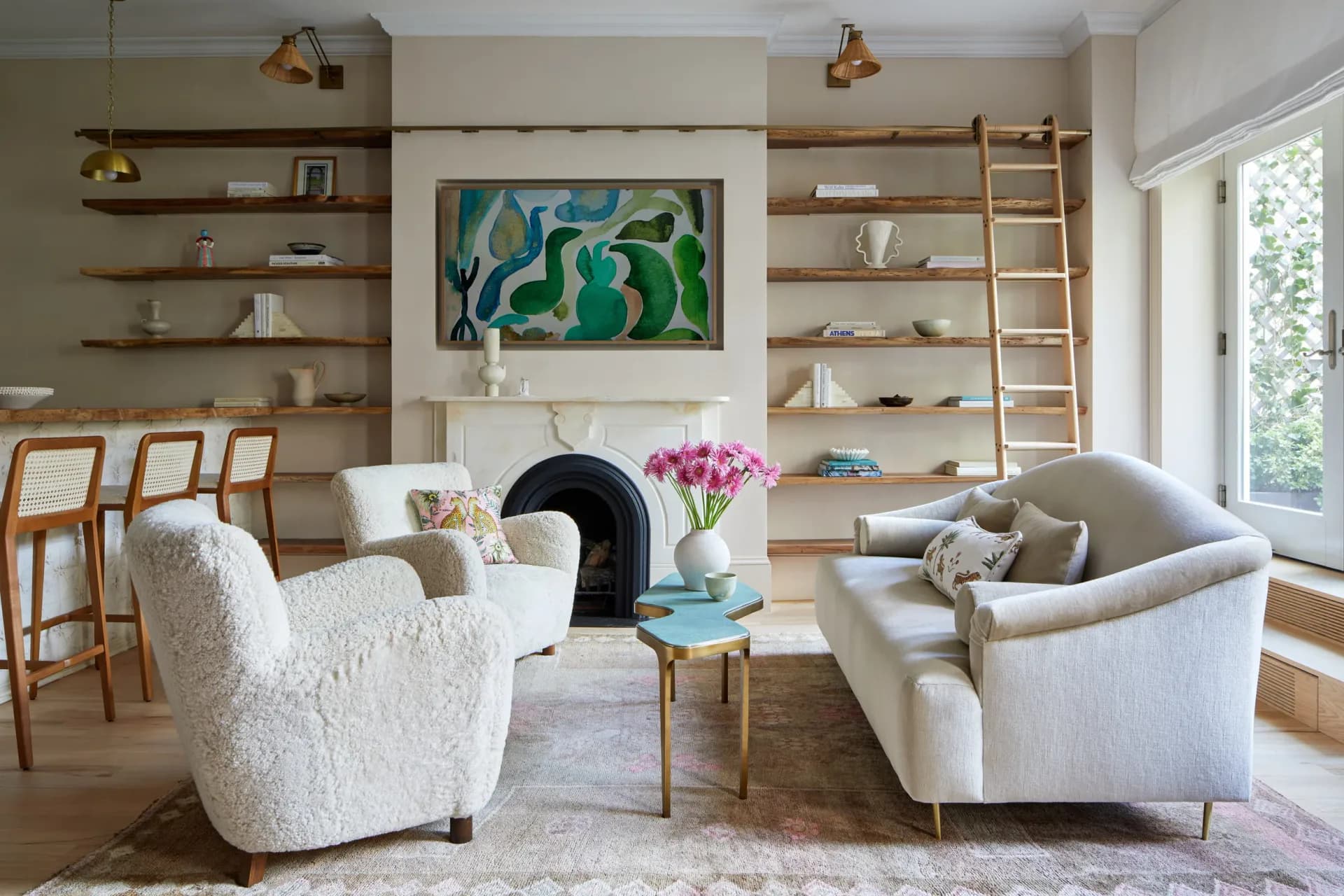 Modern living room with cream boucle chairs, wooden floating shelves, abstract green artwork, and natural light from large windows by Molly Portnof.