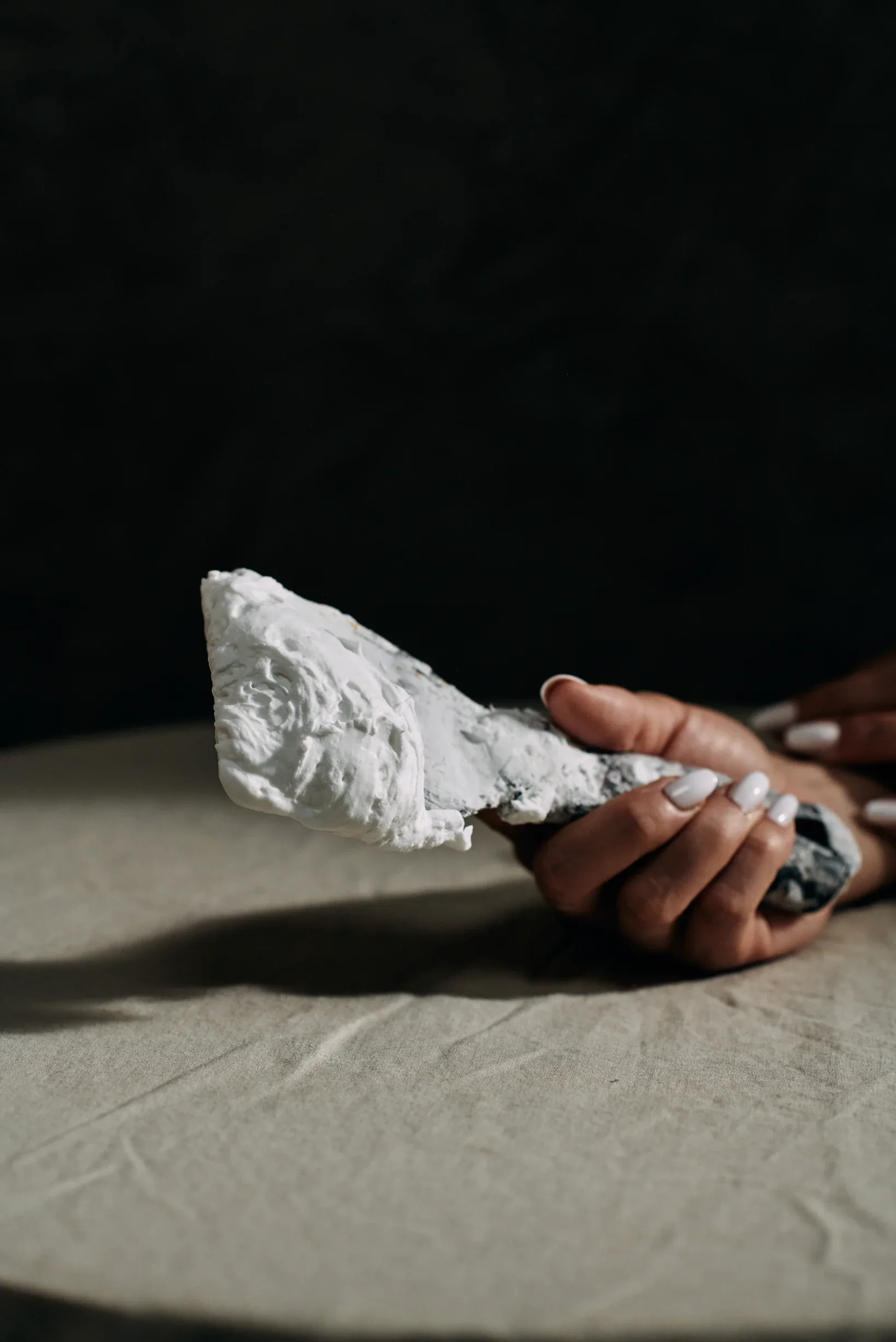 Hand holding white plaster or clay sculptural form on neutral surface with dramatic lighting by Monica Curiel.
