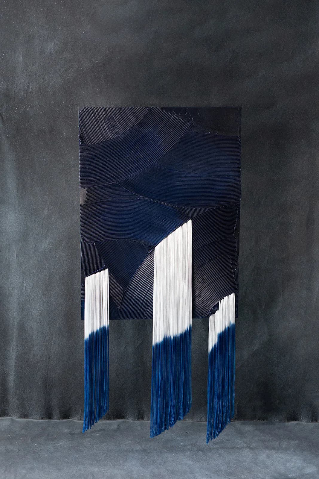 Contemporary textile installation featuring three vertical blue and white fringed panels suspended against a dark textured wall backdrop by Monica Curiel.