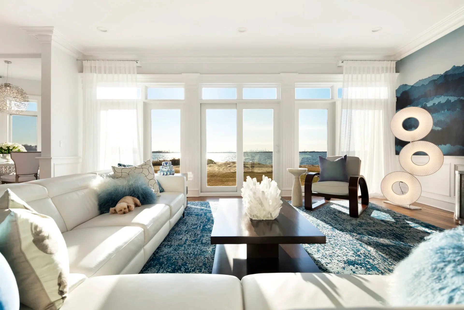 Bright coastal living room with cream sofa, black coffee table, blue area rug, and floor-to-ceiling windows overlooking water by Monica Lederman.