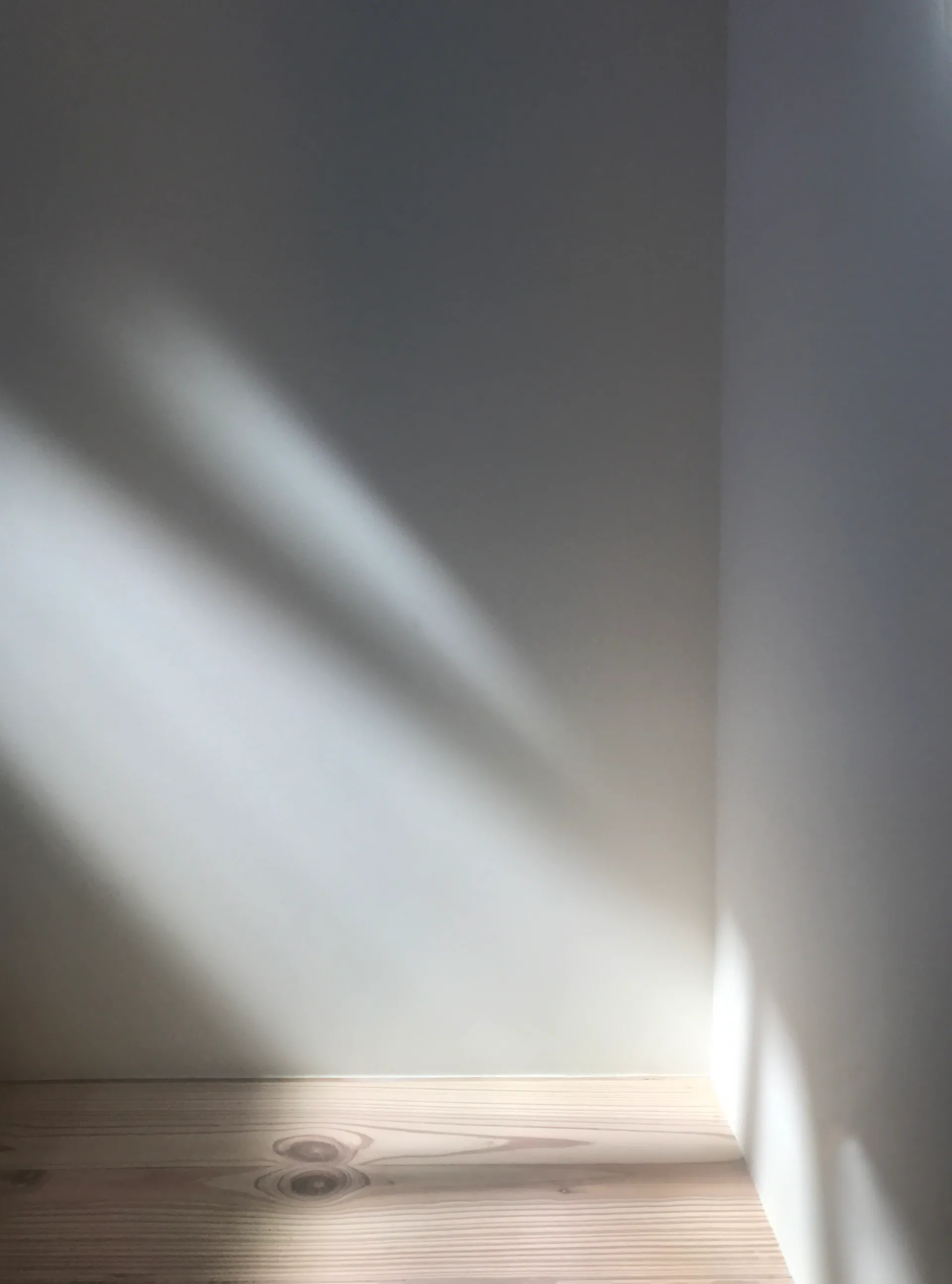 Minimalist interior with natural light streaming across light wood flooring and white walls, creating dramatic shadows and illuminating the serene space by Morgan Wendelborn.