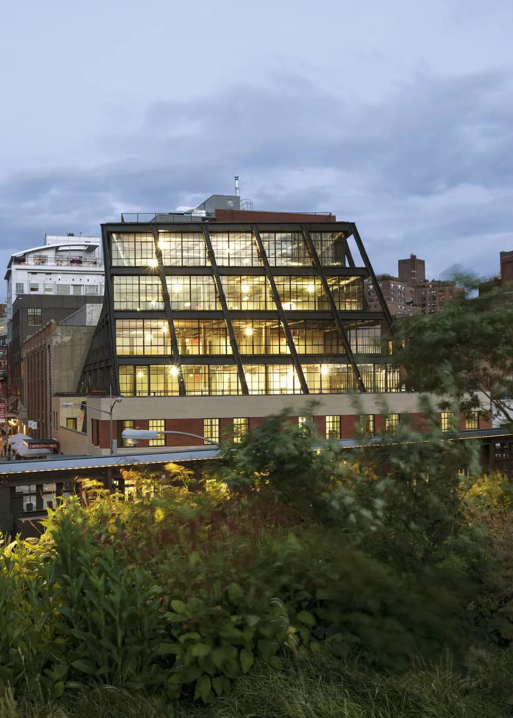 A contemporary glass-fronted building with tilted window panels and warm interior lighting integrated into a historic brick structure by Morris Adjmi.