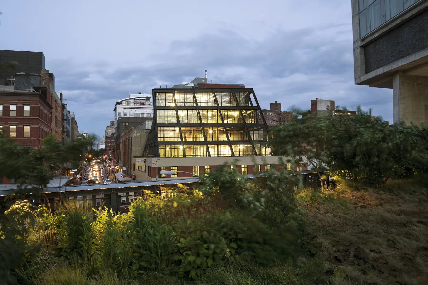 Modern angular glass and metal building with illuminated gridded facade nestled among historic brick structures and verdant plantings by Morris Adjmi.