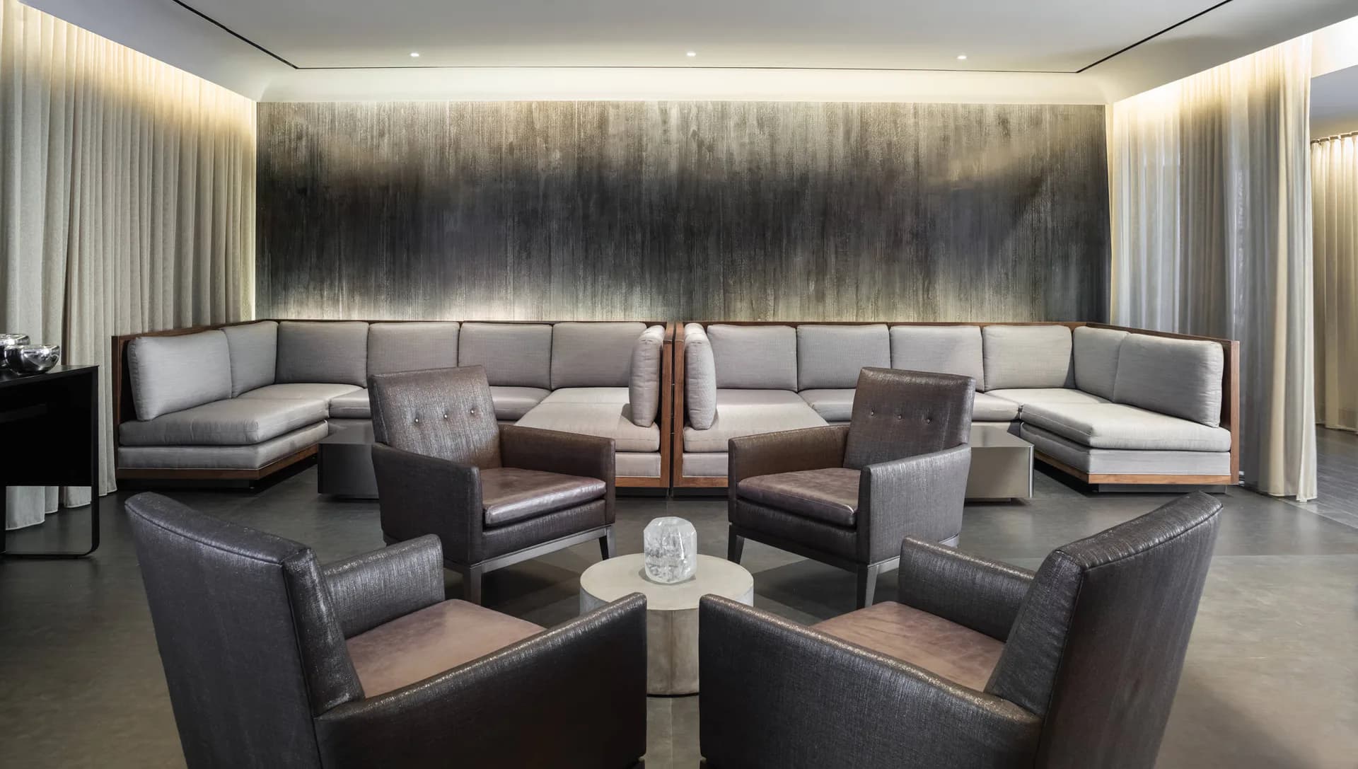 Luxurious lounge with curved grey sectional, leather armchairs, abstract wall art, and ambient lighting by Nagham Hamid.