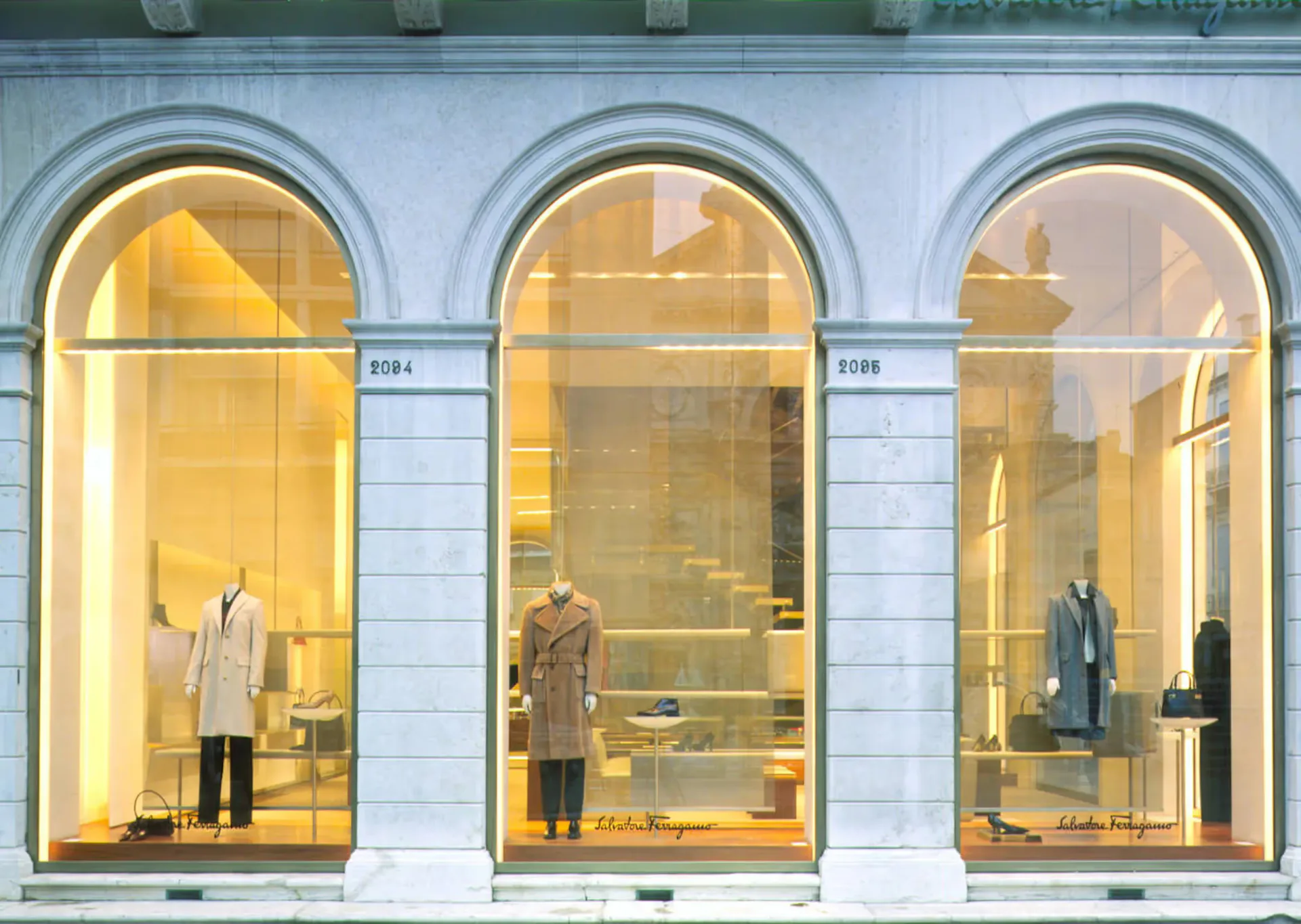 Three arched storefront windows with warm golden lighting display luxury fashion mannequins and apparel against white stone facade by Nagham Hamid.