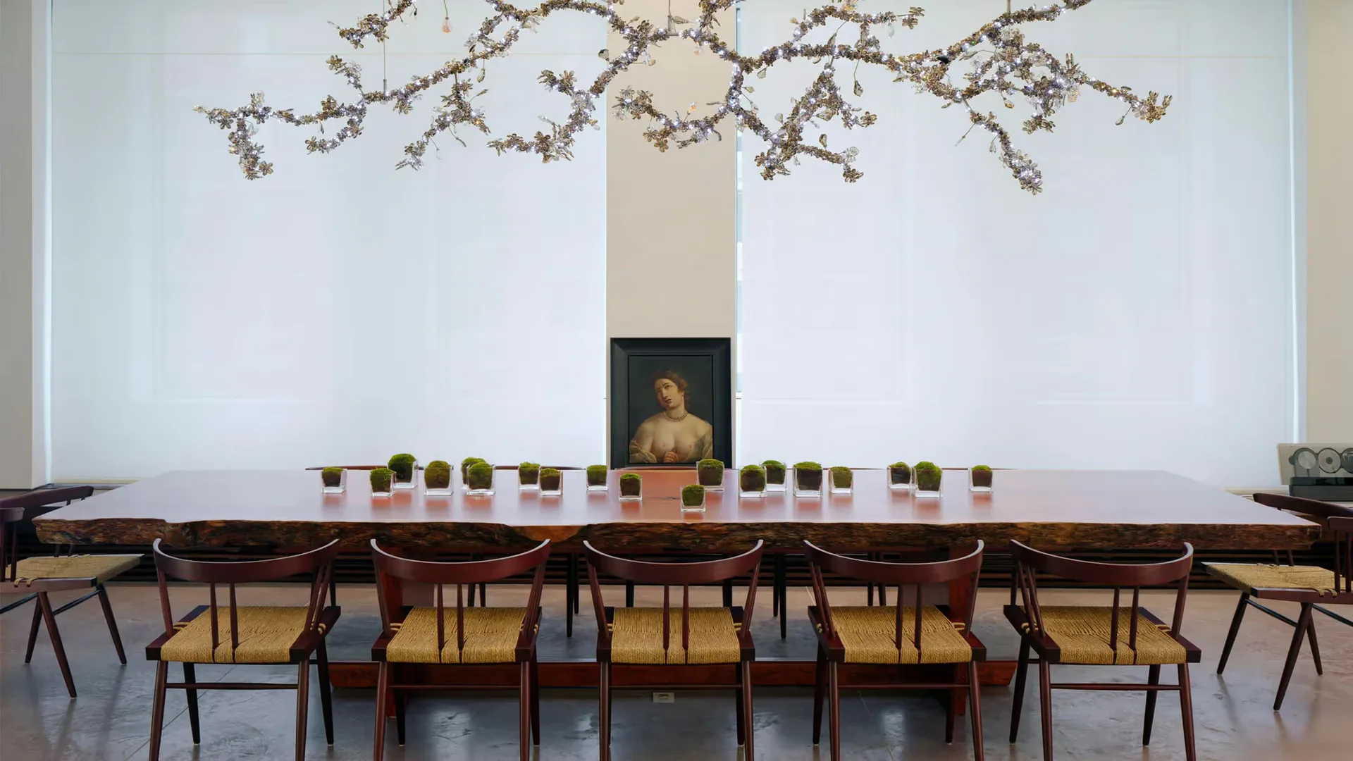 Minimalist dining room with wooden table, mid-century chairs, and dried branches overhead installation by Nagham Hamid.