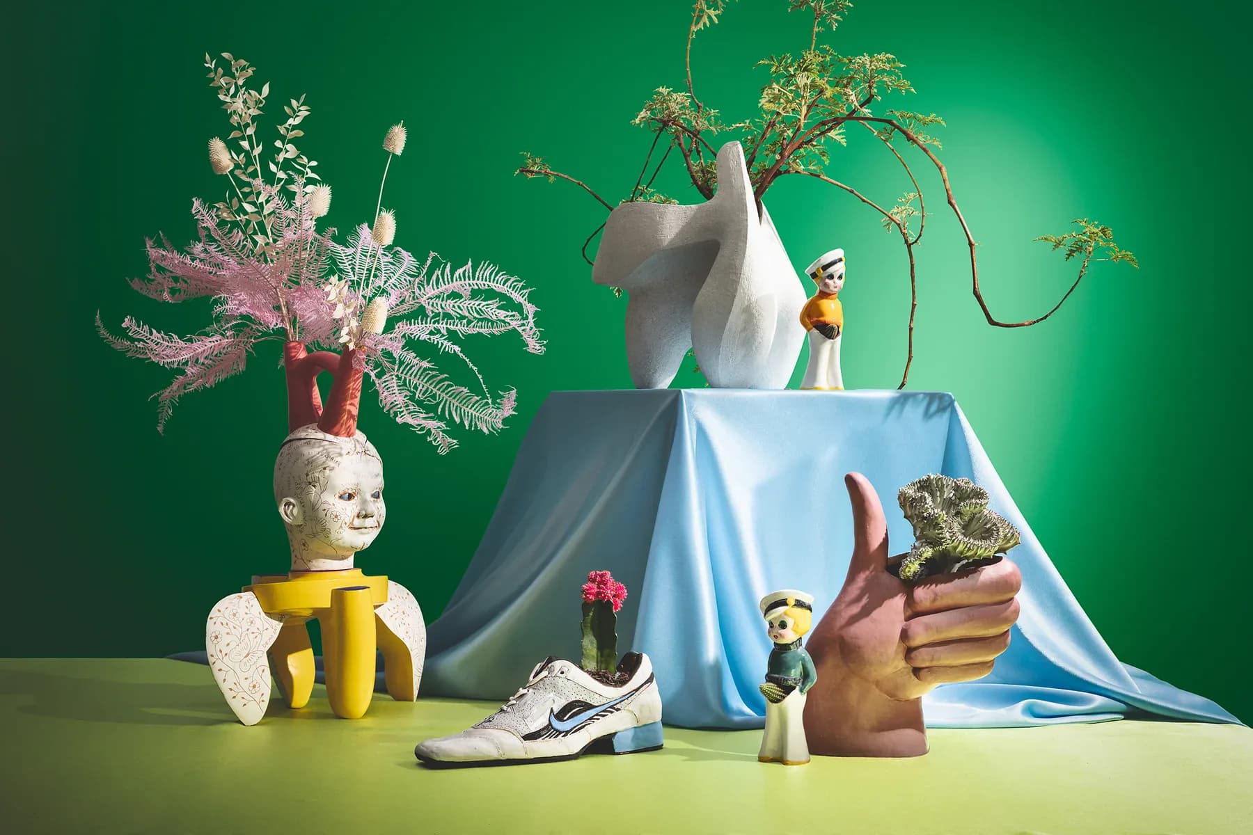 Whimsical tabletop scene with sculptural objects, potted plants, ceramic figures, and draped blue fabric against a green background by Nana Spears.