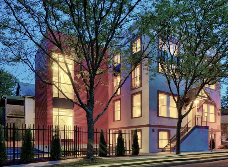 Modern residential building with warm brick and stone facades, illuminated interior windows, mature trees, and contemporary architectural detailing by Nancie Giardina.